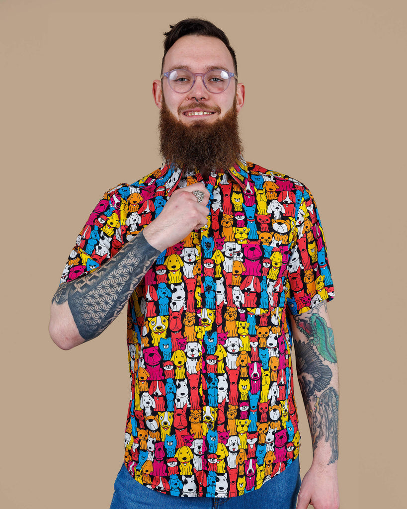 Declan a white bearded tattooed man in his 20's with brown hair and glasses is smiling at camera wearing a multicoloured shirt with dogs on and blue jeans on a beige backdrop shot in studio