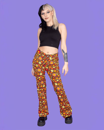 Model is wearing Ditsy Floral Bell Bottom Flares paired with a black crop top and black boots. The flares are orange and covered in red, orange and yellow flowers. The model is posing in front of a purple background facing the camera.