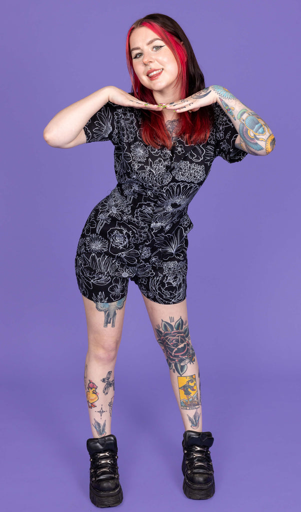 The Black Floral Stretch Twill Playsuit worn by a femme model with mid length red and black hair with black platform trainers on a purple studio background. They are facing forward smiling leaning forward with their hands framing underneath their face. The black base playsuit has white floral outlines all over.