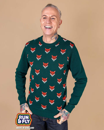 a white male tattooed model is stood in front of a beige backdrop wearing Forest Green Fox Head Jumper paired with jeans. The green long sleeved jumper has an all over print of fox heads on the torso. The model is smiling whilst holding the bottom of the jumper. 