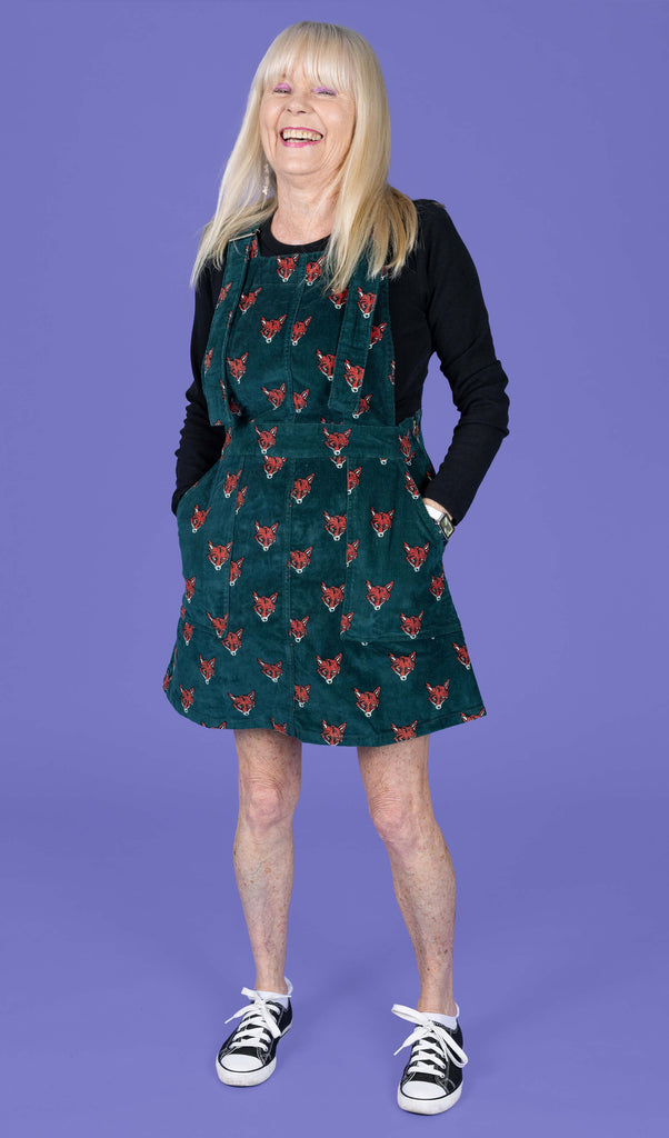 Trish, a femme white model with long blonde hair is stood in front of a purple backdrop in a photography studio in Hove wearing Fox Stretch Corduroy Pinafore Dress with a long sleeve black top underneath. The pinafore dress is a forrest green colour with an all over print of red and white fox heads. Trish is smiling at the camera with her hands in the dress pockets.