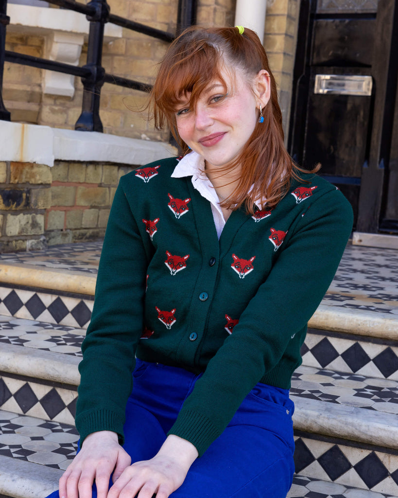 A model sitting on steps wearing a forest green cardigan with red fox head designs and blue trousers. The model has ginger hair tied with a yellow hair band and is smiling softly. The setting is outside a building with Victorian-style black-and-white tiled steps. Ethical slow fashion by Run & Fly.