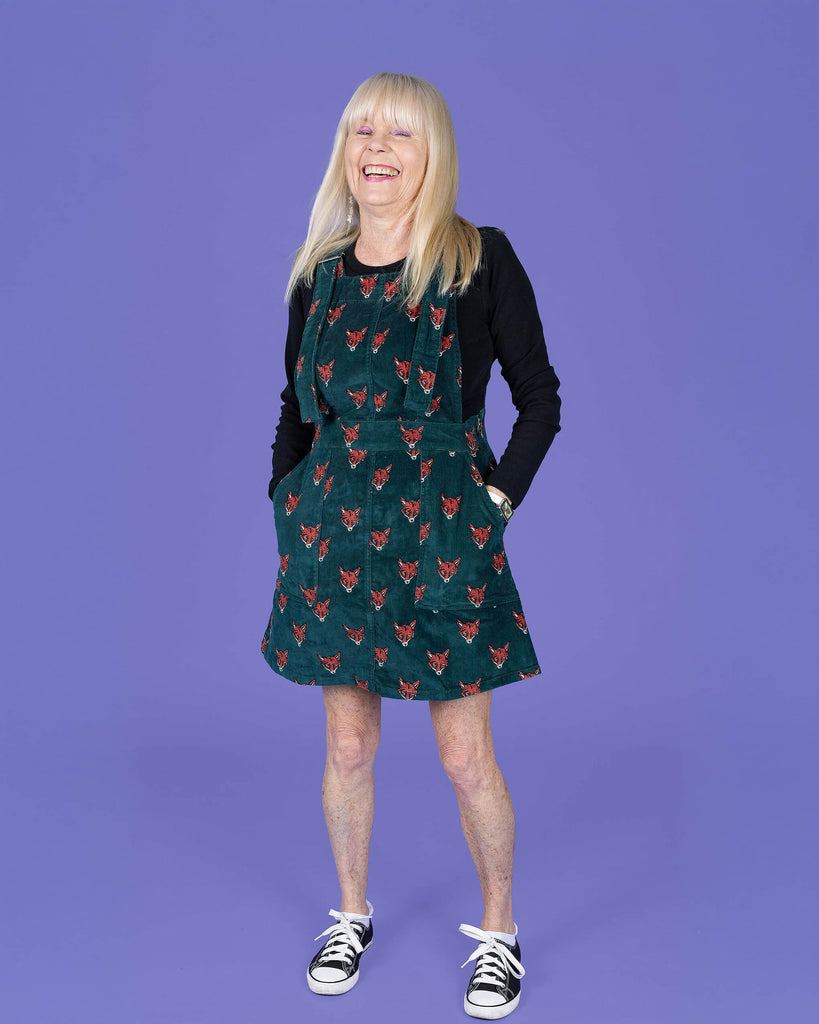 Trish, a femme white model with long blonde hair is stood in front of a purple backdrop in a photography studio in Hove wearing Fox Stretch Corduroy Pinafore Dress with a long sleeve black top underneath. The pinafore dress is a forrest green colour with an all over print of red and white fox heads. Trish is smiling at the camera with her hands in the dress pockets.