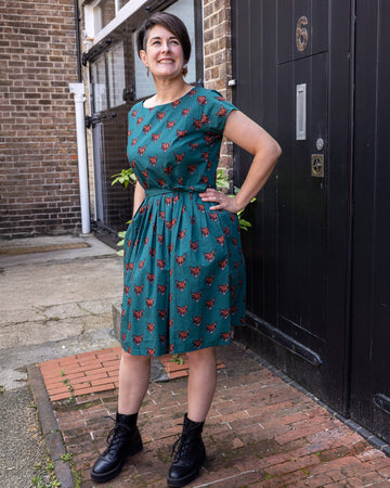 Sarah is stood by a black door outside in Hove wearing Stretch Foxy Print Tea Party Dress with Pockets with black boots. The emerald green dress has an all over fox head print. Sarah is smiling at the camera with one hand on her hip and other in the dress pocket. 