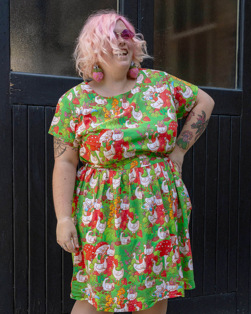 A joyful model with curly pink hair and pink sunglasses smiles while wearing a Run & Fly dress. The dress has a fun, vibrant print featuring geese and garden elements, perfect for the ethical slow fashion enthusiast. They pose against a dark wooden door.