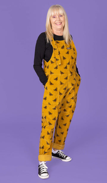 A three-quarter view of an elderly woman with long blonde hair wearing a mustard yellow corduroy dungaree with black bee patterns over a black long-sleeve shirt. She has her hands in the front pockets of the dungarees and is smiling. The background is a solid purple.