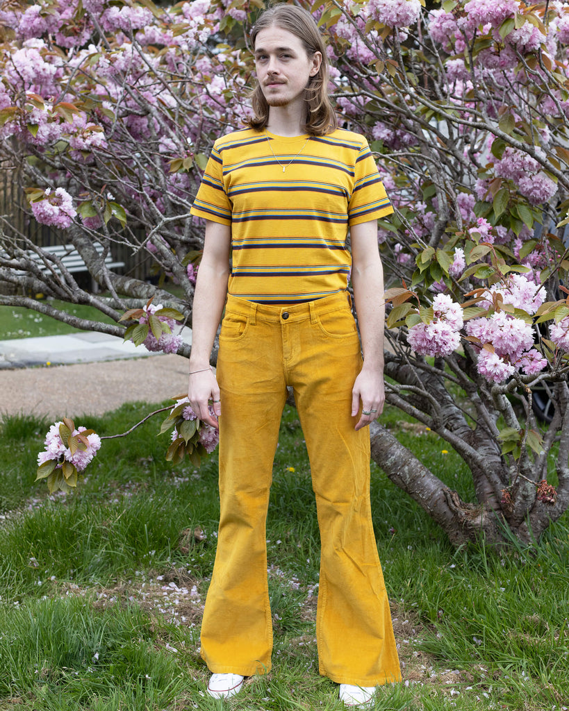 Model Jack stands confidently in Run & Fly’s Gold Corduroy Flares and a striped yellow T-shirt, surrounded by blooming cherry blossoms. The vibrant outfit adds a playful, retro vibe.