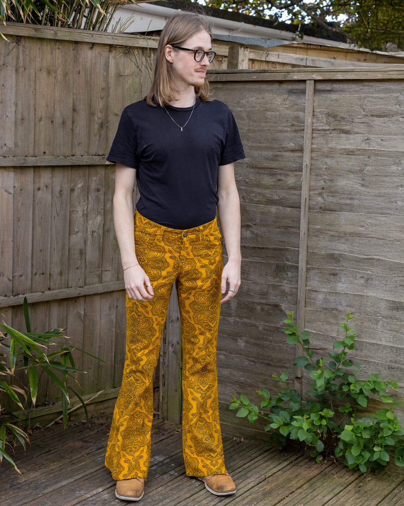 Jack, a white male model with long hair and glasses is wearing Gold Hendrix Paisley Corduroy Retro Bell Bottom Flares paired with a black t-shirt and brown shoes. The trousers are a yellow gold colour with an all over black paisley pattern. Jack is stood outside in front of a wooden fence facing the camera.