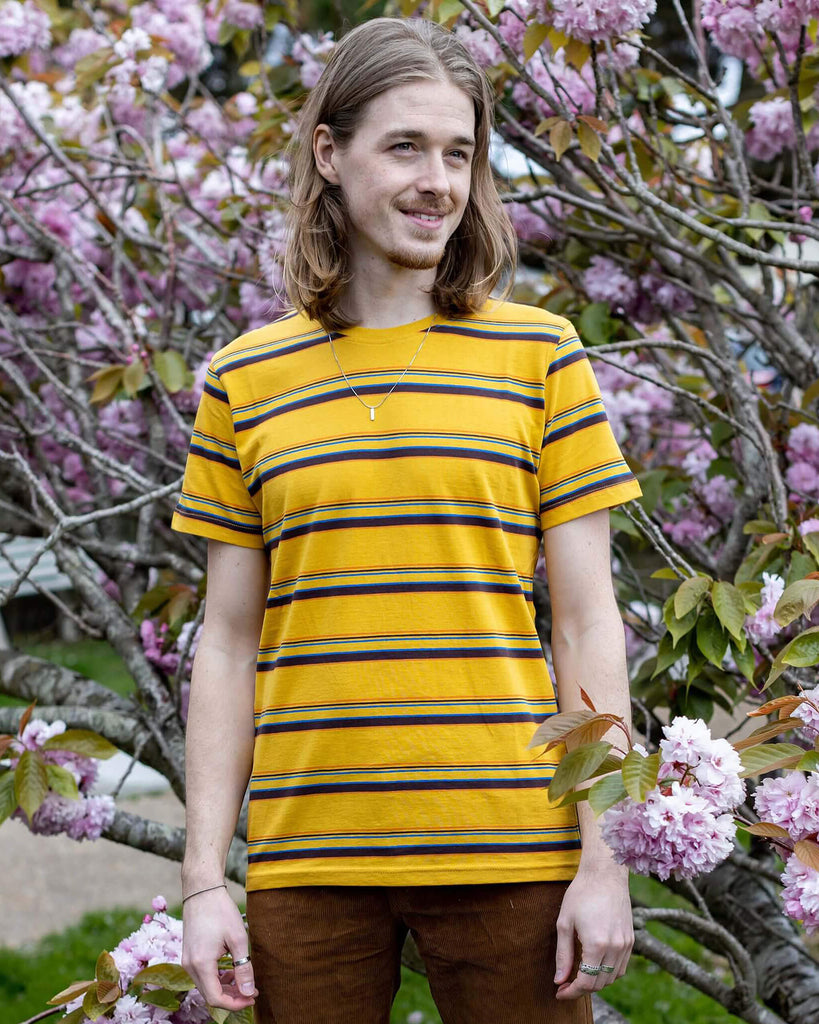 Jack is stood in front of a blossom tree wearing the gold sunset stripe retro tshirt with tobacco corduroy flares. He has mid length blonde hair and is smiling facing towards the camera with both hands resting by his sides. Photo is cropped from the knees up.