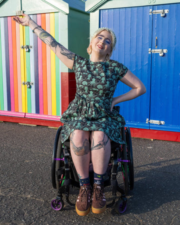 Eliza is sat outside in her wheelchair in front of beach huts on Hove seafront wearing Run & Fly Green Seas belted tea dress with pockets and brown trainers. The dress features a green octopus, squid, and nautilus print. They are posing toward the camera with one hand on their hip and other in the air. 