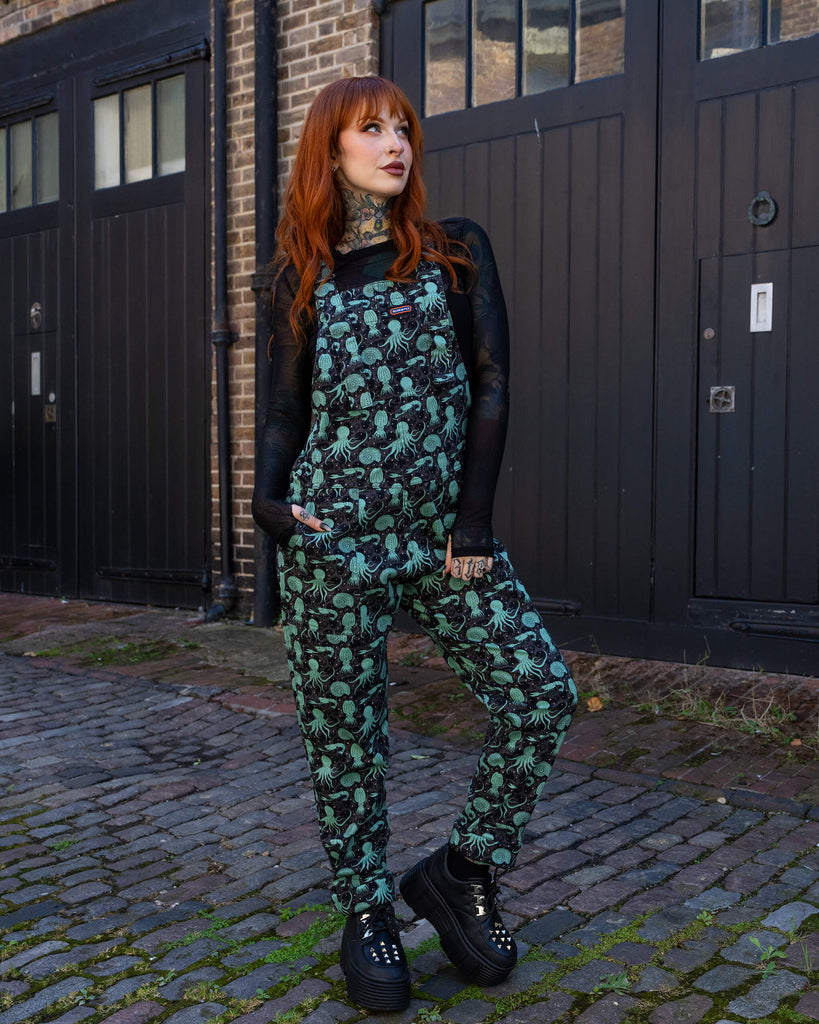 A model with tattoos and red hair stands in a relaxed pose, wearing Run & Fly black dungarees featuring a fun mint green sea creatures print. She pairs the dungarees with a black sheer top and platform boots. Run & Fly, a small ethical slow fashion brand, is known for its alternative, eco-conscious designs.