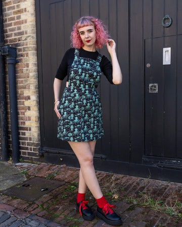 Woman wearing a black top and Run & Fly green sea creatures pinafore dress. The dress features an octopus, squid, and nautilus print. She stands outside in front of a dark wood door, posing with one hand on her pink hair and the other hand in her pocket. Ethical slow fashion brand, Run & Fly.