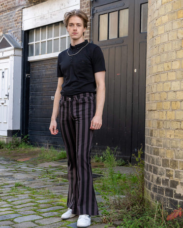 Front view of a model wearing Run & Fly grey and black vertical striped flare trousers. The trousers sit high on the waist, featuring bold stripes and a flared leg, perfect for retro or alternative looks. Paired with a simple black t-shirt, the model poses confidently in a cobblestone alley. Ethically made by a small slow fashion brand.