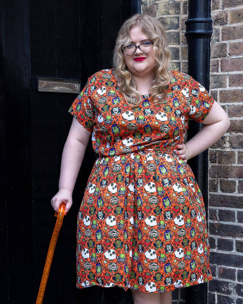 a white femme model with red lipstick wearing Halloween: Party Frogs Stretch Belted Tea Dress with Pockets. The orange dress has an all over print of cartoon frogs in various Halloween costumes such as ghosts and zombies. The model is stood outside in Hove smiling towards the camera with one hand on their hip and other holding an orange walking aid.