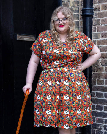 a white femme model with red lipstick wearing Halloween: Party Frogs Stretch Belted Tea Dress with Pockets. The orange dress has an all over print of cartoon frogs in various Halloween costumes such as ghosts and zombies. The model is stood outside in Hove smiling towards the camera with one hand on their hip and other holding an orange walking aid.