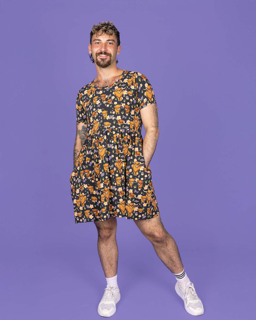 The Highland Cows Oversized Smock Dress worn by a masc non binary person with a short curly brown mullet with white trainers on a purple studio background. They are facing forward smiling leaning more on their left leg with their hands in the dress pockets. The black base dress features grass, purple and yellow flowers and brown highland cows eating grass.