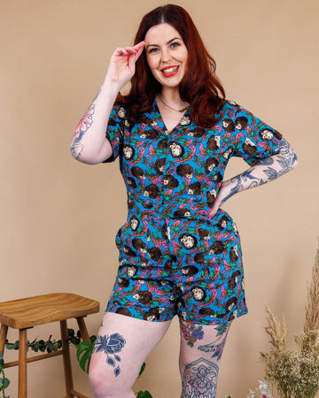 Tattooed model with red hair wearing Hedgehog Stretch Playsuit paired with lime green chunky boots. The playsuit is a blue colour with an all over print of brown hedgehogs and green, pink and orange leaves. The collared playsuit is buttoned up, with the top button left undone. Model is posing with one hand on hip and other hand in her hair.