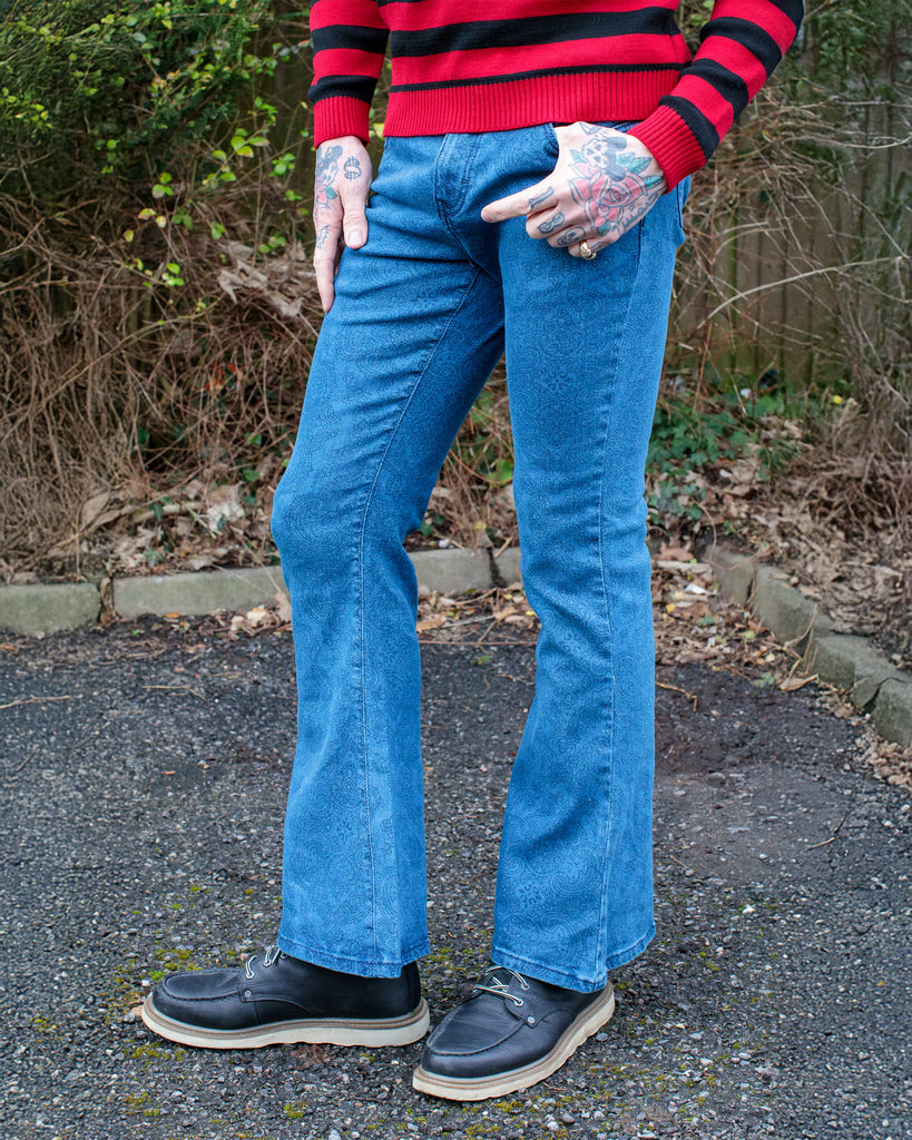 Jim is stood in a garden wearing the retro stretch stone wash denim Hendrix paisley bell bottom flares with a black and red striped jumper and black boat shoes. They are facing forward posing with one leg bent forward to highlight the flare shape.