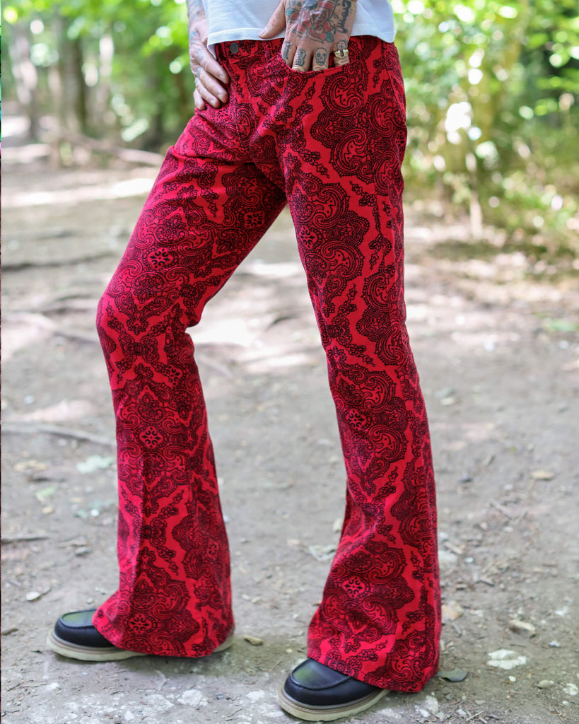 Model is stood in a wooded area wearing the Hendrix paisley corduroy retro bell bottom flares with a white short sleeve tshirt and brown boat shoes. They are facing to the left and posing with one leg out to the side to highlight the flares. Photo has been cropped from the waist down.