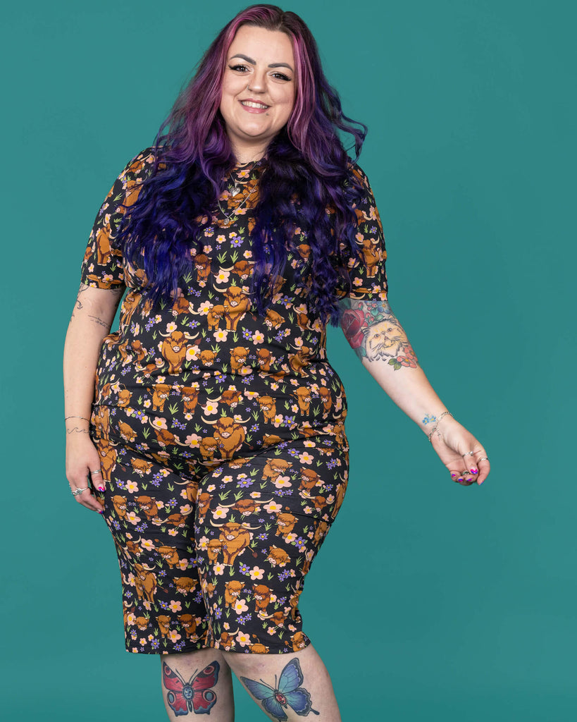 The Highland Cows Stretch Cycle Shorts worn by a femme alternative tattooed model with long pink and purple hair, the matching tshirt and black boots on a teal studio background. She is facing forward smiling leaning slightly back with one leg bent and their arm out to the side. The black base shorts features grass, purple and yellow flowers and brown highland cows eating grass.