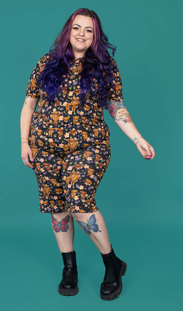 The Highland Cows Stretch Cycle Shorts worn by a femme alternative tattooed model with long pink and purple hair, the matching tshirt and black boots on a teal studio background. She is facing forward smiling leaning slightly back with one leg bent and their arm out to the side. The black base shorts features grass, purple and yellow flowers and brown highland cows eating grass.
