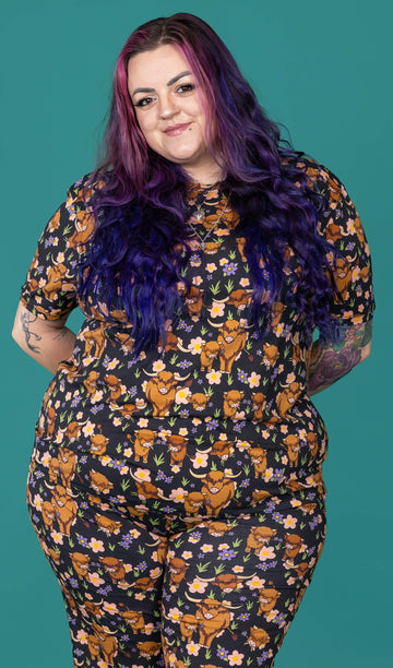 The Highland Cows Short Sleeve T Shirt worn by a femme alternative tattooed model with long pink and purple hair, the matching cycle shorts on a teal studio background. She is facing forward smiling with her arms resting behind her back. The black base t shirt features grass, purple and yellow flowers and brown highland cows eating grass.