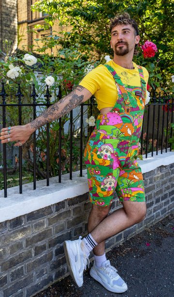 The Run & Fly x The Mushroom Babes Hills Stretch Twill Dungaree Shorts worn by a masc non binary person with a curly brown mullet haircut, a yellow crop top and white trainers. They are stood outside in front of a black railing with foliage and rose bushes behind. They are facing forward leaning off the railing with one leg crossed in front of the other smiling looking left. The dungashorts are green, pink and orange hills with mushroom houses, flowers, rainbows and trees.