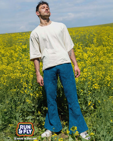 Zach is stood in a yellow flower field wearing the ink blue paisley corduroy bell bottom super flares with an oversized cream tshirt and white trainers. He is leaning back with his eyes closed.