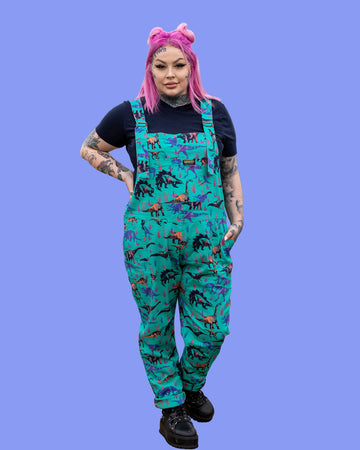 Zoe wearing the jade adventure dinosaur stretch twill dungarees with a short sleeve black tshirt and black boots. She is facing forward standing on a purple background posing with one arm on her hip and the other in the dungaree pocket.