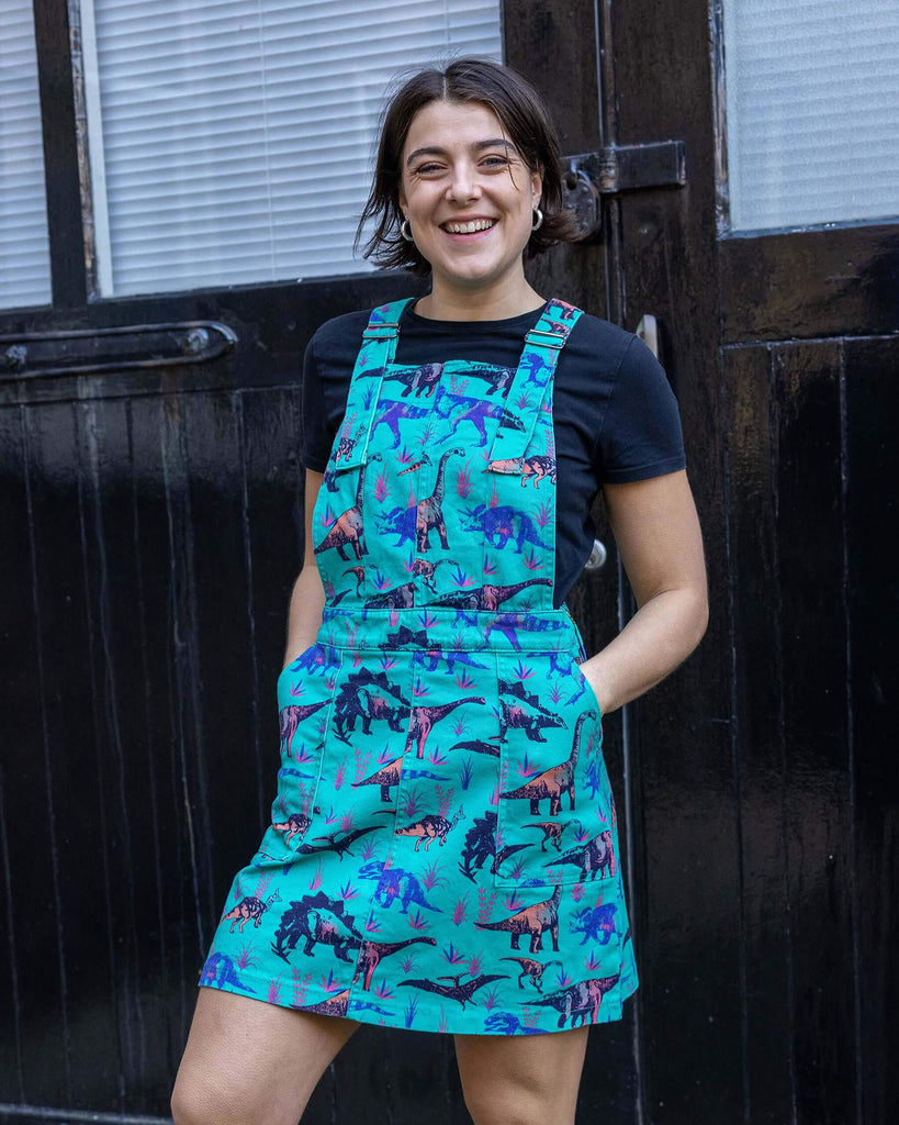 Freya is wearing the blue Dinosaur stretch twill pinafore dress, worn with a black short sleeved top and chunky boots. Model faces camera with hands in pockets, smiling in front of a garage door.