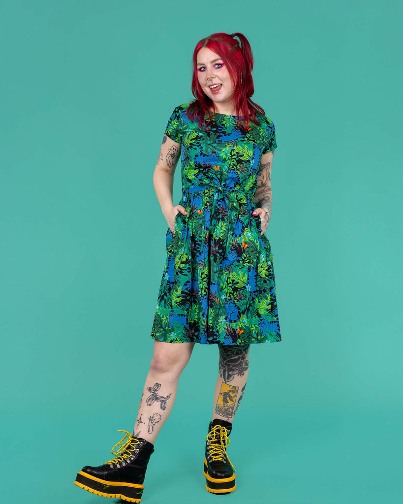 The Jungle Cats Stretch Belted Tea Dress with Pockets being worn by Flo on a teal background. Print features various green, brown and black foliage with orange butterflies and dark blue jumping tabby cats, the dress is short sleeve with a belt tie and pockets. Flo has red hair in bunches, neon pink makeup and tattoos with yellow and black platform boots. She is facing forward smiling leaning back on one leg with both hands in the dress pockets.