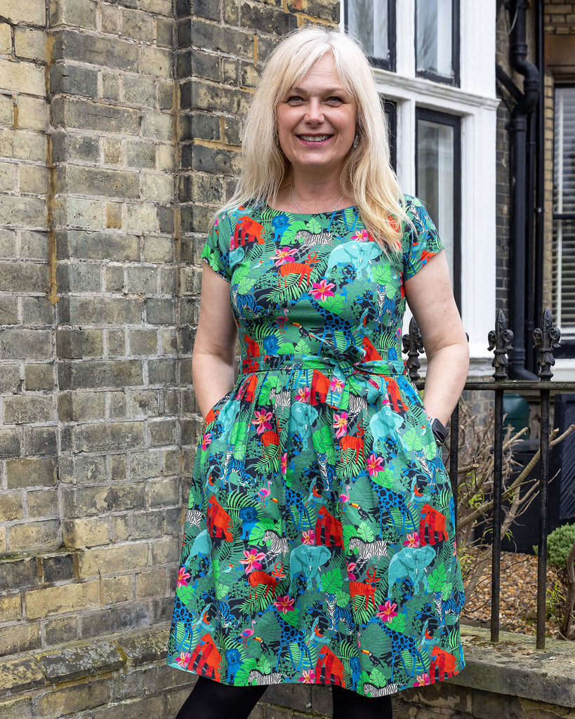 Front view of a blonde woman wearing the vibrant Jungle Stretch Belted Tea Dress by Run & Fly, a small ethical slow fashion brand. The dress features a lively print of jungle animals, including tigers, elephants, and giraffes, with a belted waist and pockets. She stands in front of a brick wall, smiling warmly.