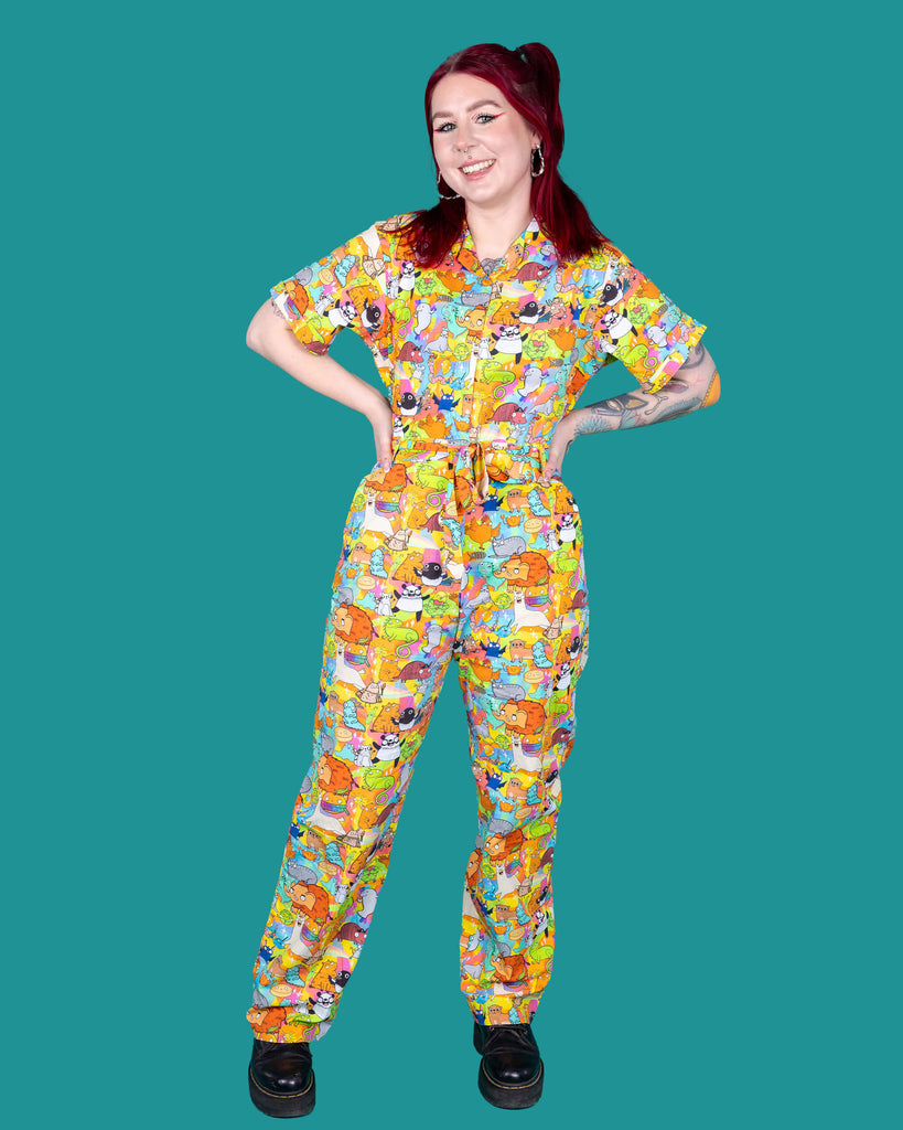 Florence a red haired female model with tattoos and cool make up is wearing a colourful rainbow jumpsuit with cute drawing by Katie Abey. She is standing against a turquoise background and smiling at camera. 