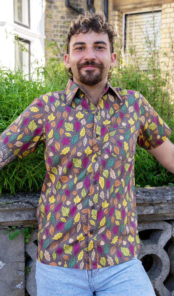 The Autumn Leaves Short Sleeve Shirt worn by a masc non binary person with a curly brown mullet cut and denim shorts. They are stood outside along the pavement in front of a bush and ornate wall facing forward smiling leaning back on the wall. The light brown based shirt features dark brown, yellow, orange, dark red, dark green and beige leaves with acorns all over. 