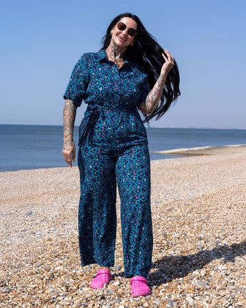 The Atlantic Blue Leopard Print Jumpsuit worn by a femme tattooed model with long black hair wearing pink crocs shoes and sunglasses. She is stood outside on a sunny pebble beach with the sea in the background. She is facing forward laughing brushing one hand through her hair. The blue base jumpsuit features pastel blue, lilac, purple, dark blue and black leopard print spots.