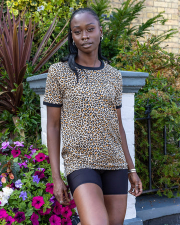 black femme model with piercings is stood outside in Hove by plants wearing Natural Leopard Print T Shirt paired with black shorts. The model is posing towards the camera with arms by her side. 