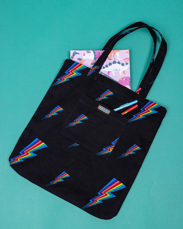 a black tote bag with all over rainbow lightning bold print. The tote bag is laid flat on a green background with a book in the main section of the bag the smaller front pocket has Run & Fly logo on the front.