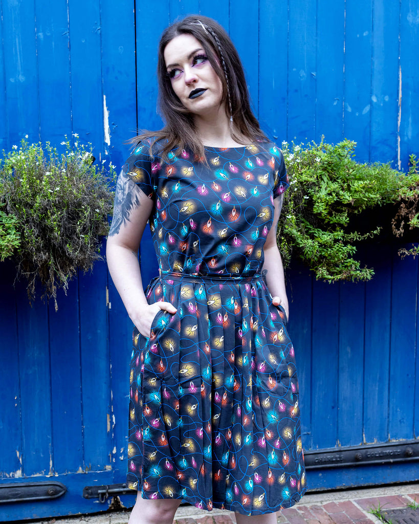 Alice a brown haired tattooed model wearing a fairy lights tea dress by Run & Fly against a blue door. The print is a blue base with multicoloured fairy light bulbs glowing all over.