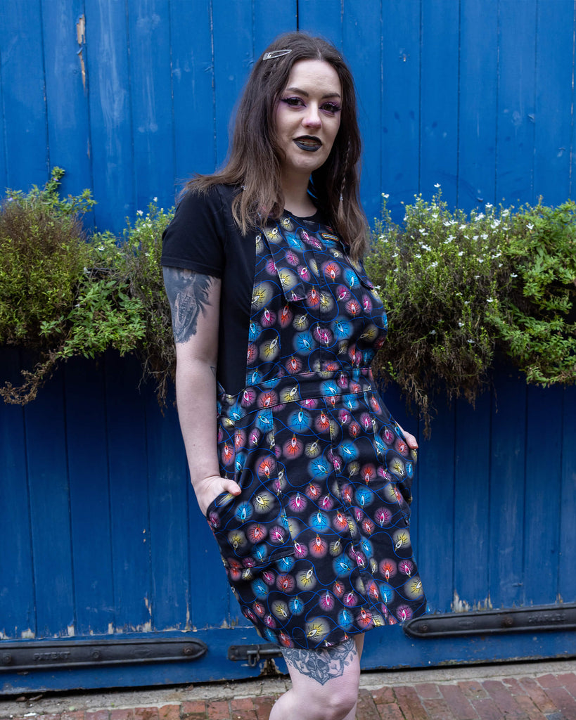 Alice a brown haired tattooed model wearing a fairy lights pinafore  dress by Run & Fly against a blue door. The print is a blue base with multicoloured fairy light bulbs glowing all over.