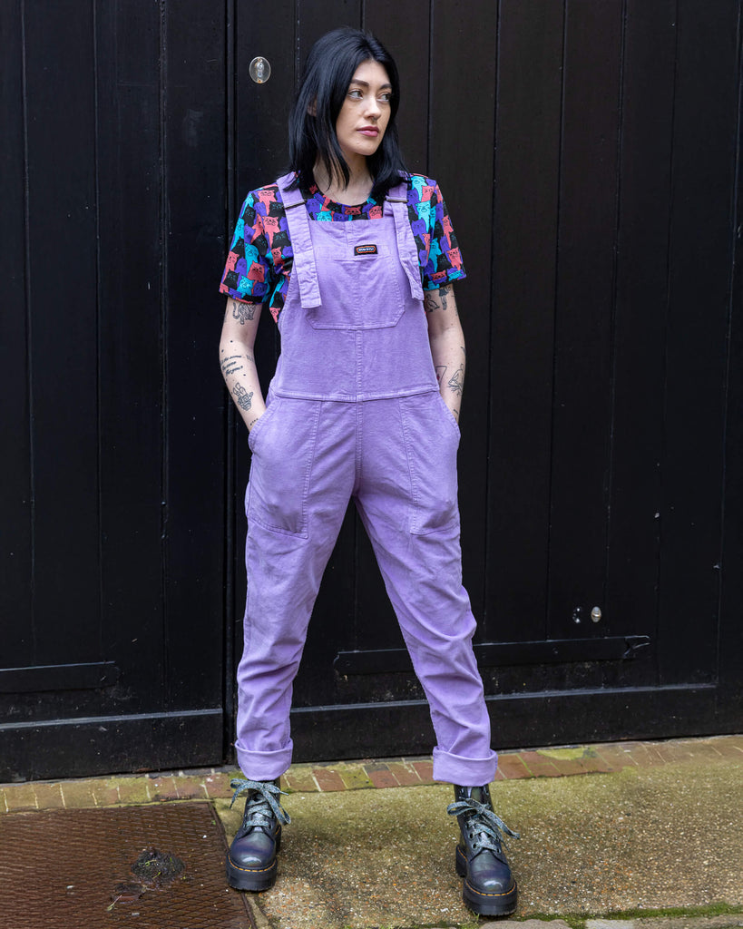 white femme model with black hair and tattoos is stood in Hove in front of a pink background wearing Lavender Stretch Corduroy Dungarees paired with Cat Chorus Short Sleeve Tee and silver boots. The model is posing facing the camera with one hand in the dungaree pocket and other tucking her hair behind her ear.  