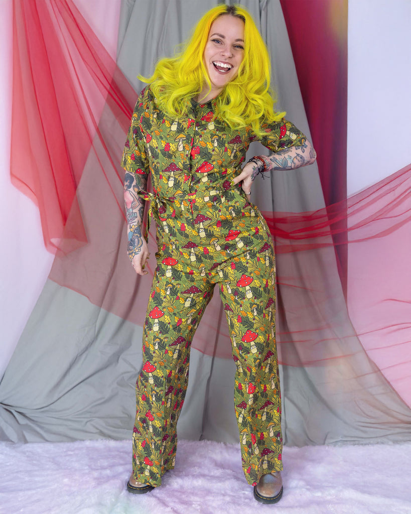 Katie Abey is stood in a photography studio in Hove wearing Run & Fly x Katie Abey Mushroom Boi's Jumpsuit. The jumpsuit has an all over 'mushroom bois' print illustrated by Katie Abey. Katie is smiling and facing the camera with her hand on her hip.