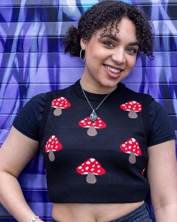  Georgia is stood in front of a graffiti wall wearing the mushroom knitted tank top over a black short sleeve shirt and black jeans. She's posing facing the camera smiling with one hand in the front jean pocket. Photo is cropped from the hips up.