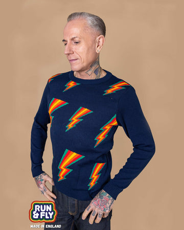 James is stood in front of a beige background wearing the run and fly navy lightning bolt jumper with denim jeans. He is facing left with both hands on his hips whilst looking down. The jumper is a navy blue base colour with repeating green, red, orange and yellow lightning bolts on the chest. The arms and back are plain navy blue. The photo is cropped from the hips up. In the left corner is a Made in England logo.