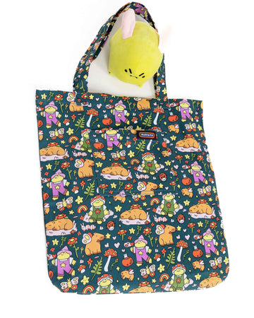 The Run & Fly x Nimbob Capybara Fairies Tote Bag laying flat on a white background with the nimbob grumpy frog plush. The green base tote bag feature frogs in dungarees, butterflies, plants, potion bottles, mushrooms and capybaras with fairy wings.