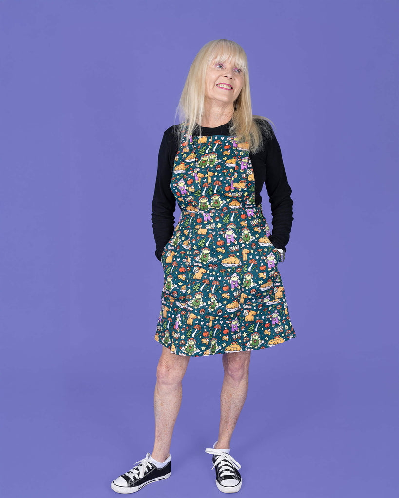 The Run & Fly x Nimbob Capybara Fairies Stretch Twill Pinafore Dress worn by a femme blonde lady with a black long sleeve tshirt and black converse style trainers on a purple studio background. She is facing forward smiling looking off to the right with both hands resting in the dress pockets. The green base pinafore feature frogs in dungarees, butterflies, plants and capybaras with fairy wings.