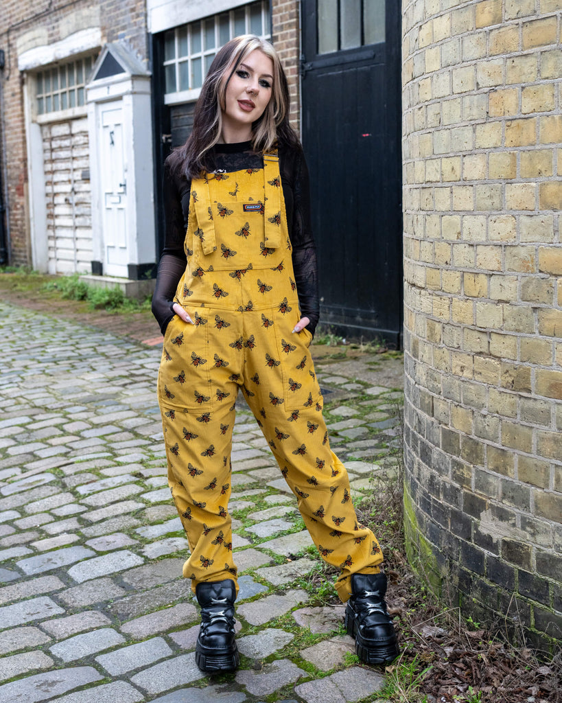 A model poses on a cobbled street wearing Run & Fly's Gold Bee Corduroy DungaBee Dungarees, a mustard yellow corduroy design with an all-over black and orange bee print. They pair the dungarees with a black mesh top and chunky black platform boots. Their hands are tucked into the pockets as they lean casually against a brick wall.