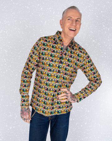 James, a tattooed male model, is smiling brightly while wearing a fitted long-sleeve button-up shirt with a vibrant Nutcracker print designed by Waffle Mama Design. The Nutcrackers feature bold and colourful tones of red, yellow, green, and blue. The snowy background complements the festive aesthetic.