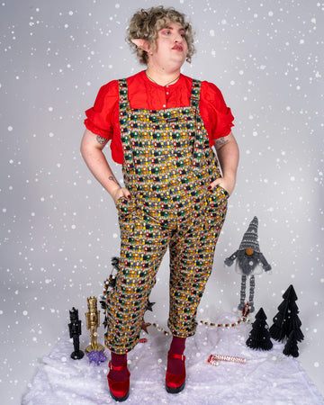 Model Maddie, wearing Run & Fly’s Nutcracker print dungarees in a festive setting, poses with joy. The colourful print features a variety of nutcrackers in vivid outfits on a dark background. Maddie pairs the look with a red blouse, red velvet shoes, and maroon tights, adding a holiday flair. The scene includes holiday decorations like a mini Christmas tree and gnome, enhancing the festive vibe.”