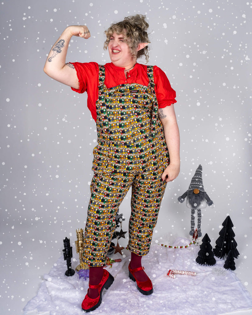 In a playful pose, model Maddie shows off Run & Fly’s Nutcracker print dungarees. The colourful nutcracker print contrasts beautifully with the red blouse and festive accessories, including maroon tights and red shoes. Holiday decorations, such as a gnome and Christmas ornaments, surround her in a wintery scene.