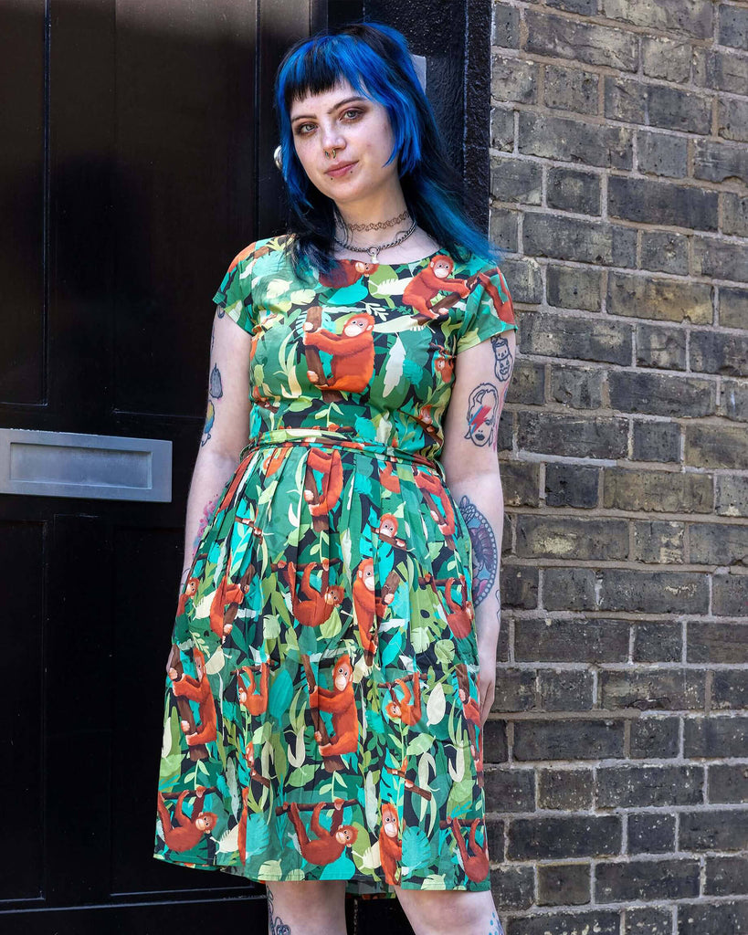 Faeryn is stood outside in Hove in a mews wearing Orangutan Stretch Belted Tea Dress with Pockets paired with black boots. The green dress has an all over orangutan print with leaves and branches. Faeryn is posing facing the camera. 